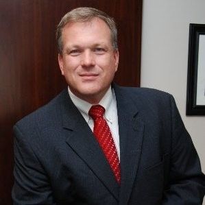 Smiling man in a navy blazer and checkered shirt, standing in an office setting with blurred background