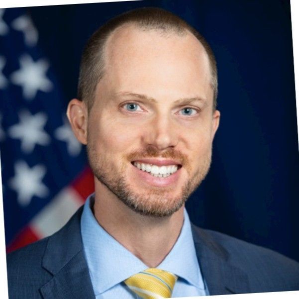 Smiling man in a navy blazer and checkered shirt, standing in an office setting with blurred background