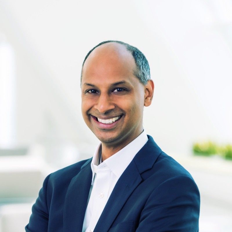 Headshot of a smiling person in a dark suit and white shirt on a gray background