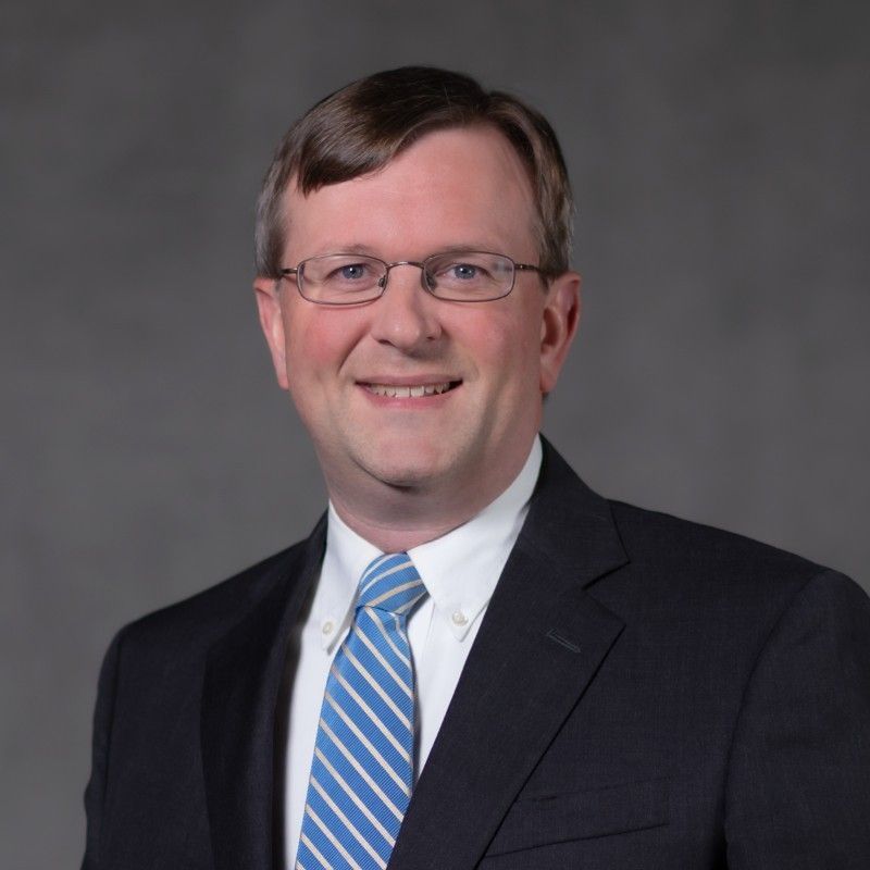 Headshot of a smiling person in a dark suit and white shirt on a gray background