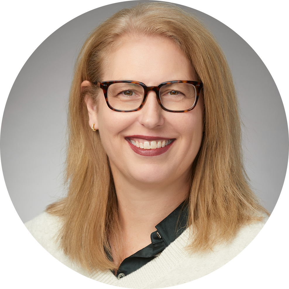Smiling woman with blonde hair and glasses, wearing a white sweater and dark blouse on a gray background
