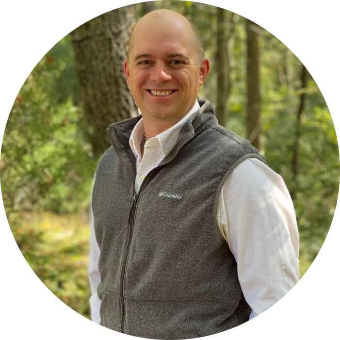 Smiling man in a gray vest and white shirt standing in a wooded outdoor setting