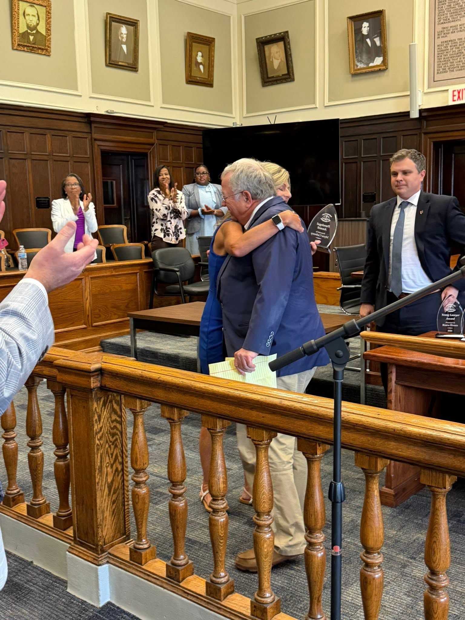 Two people embrace in a wood-paneled courtroom while others stand nearby.