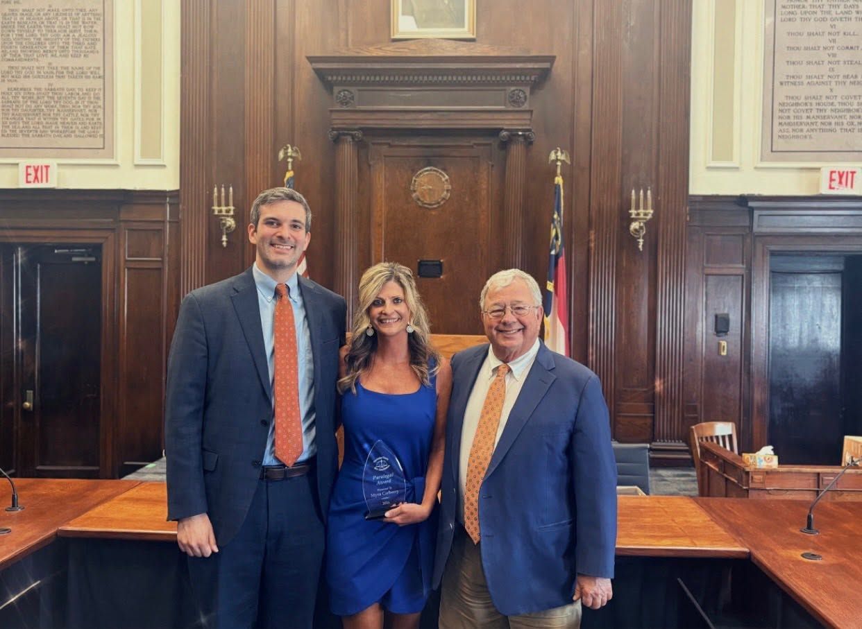 Three people posing in a wood-paneled courtroom, smiling in front of the judge’s bench.