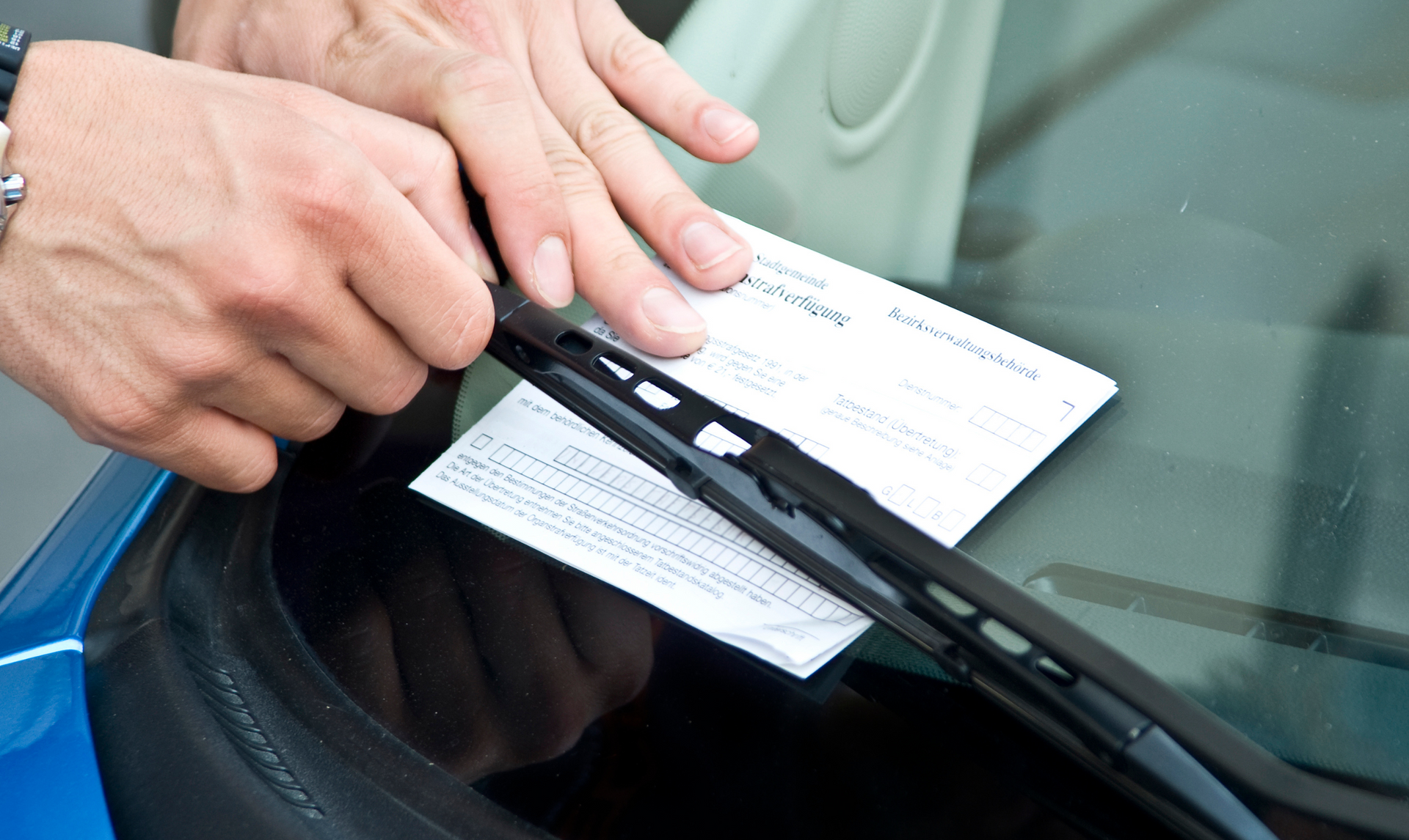 Hands placing a parking ticket under a windshield wiper on a blue car.