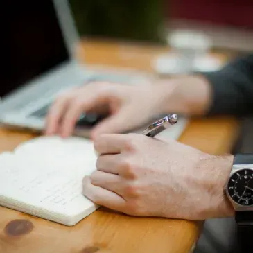 A person is writing in a notebook while using a laptop computer.
