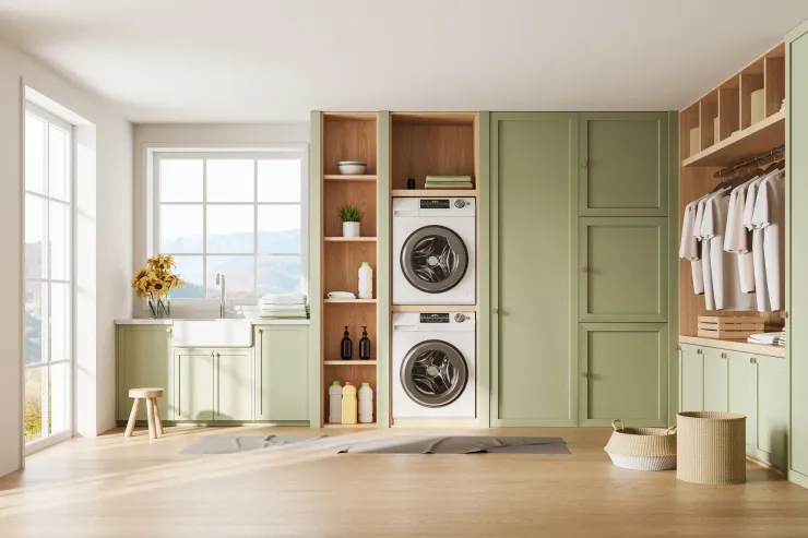 Bright laundry room with stacked washer-dryers, sage cabinets, shelving, and a window-lit sink.