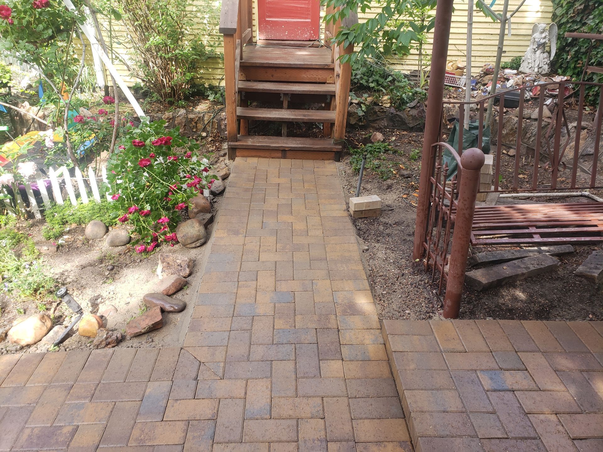Brick garden path leading to wooden steps and a red door, with flower beds and a fenced side area