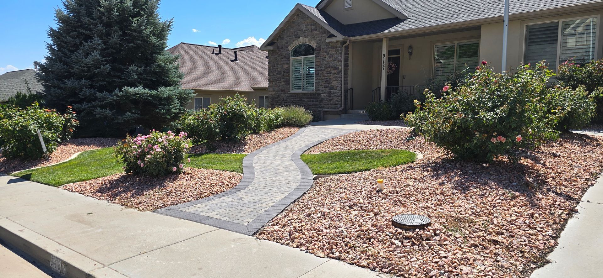 Front yard landscaping with a curved concrete walkway, mulch beds, shrubs, and a brick house in the background