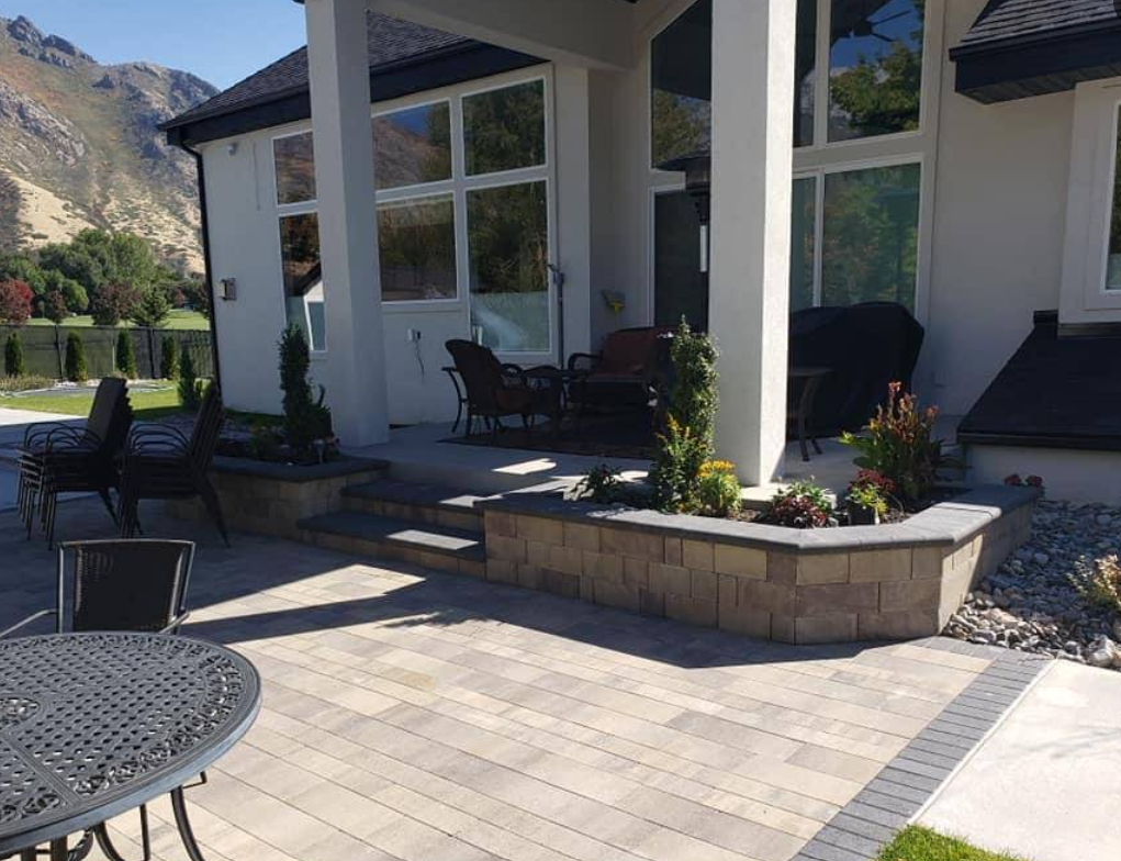 Modern patio with stone pavers, black outdoor furniture, and a white house porch with planters and mountains nearby