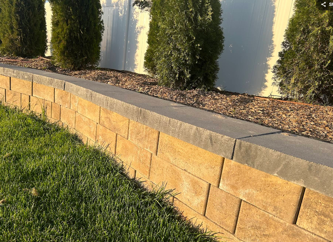 Curved tan stone retaining wall beside grass and shrubs in a landscaped yard