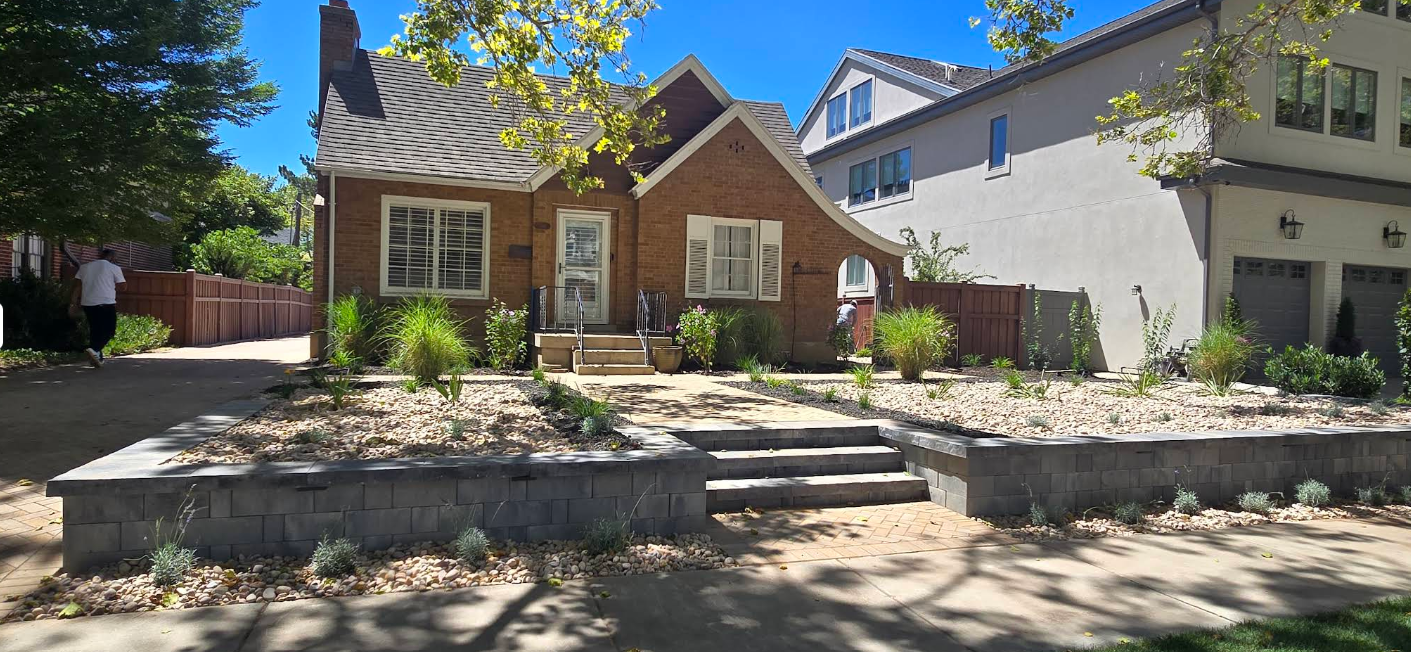 Small brick house with a front garden, stone retaining wall, and a tree in a sunny residential neighborhood