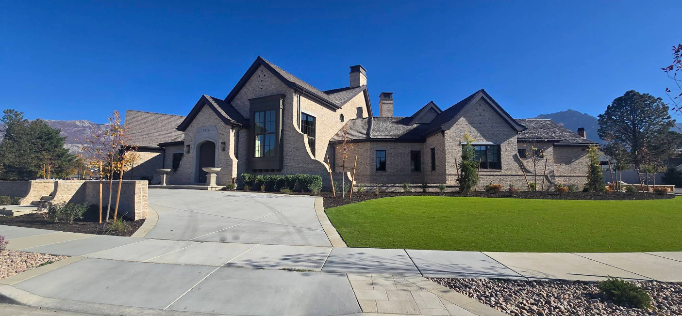 Large beige stone house with a steep roof, driveway, and manicured green lawn under a clear blue sky