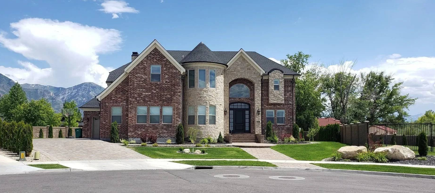 Large stone suburban house with arched entry, driveway, and manicured lawn under a blue sky