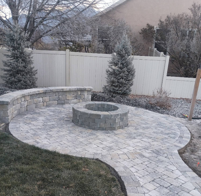 Backyard patio with stone fire pit, curved retaining wall, paver path, and white fence in winter.