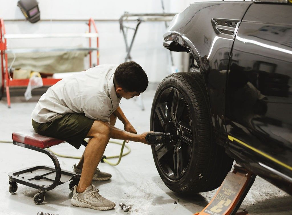 Inside an automotive workshop, a technician is using a drill to remove the lug nuts on the tire of a