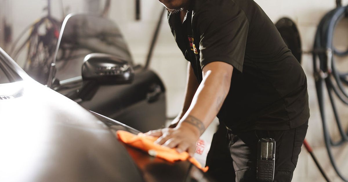 A man wiping the front of a black car with an orange cloth inside an automotive service area with eq