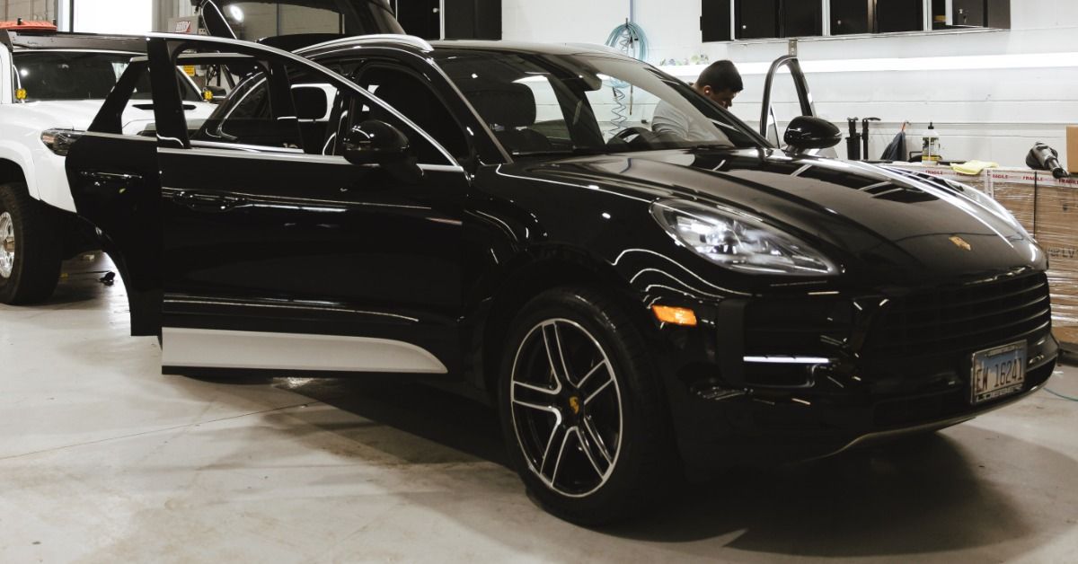 A black car with open doors parked inside a service bay beside another vehicle, boxes, tools, and wall cabinets.