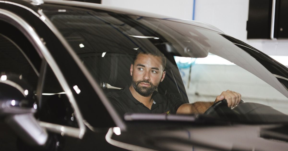 A man sitting behind the wheel of a black car inside a service bay with bright lights reflecting on the windshield.