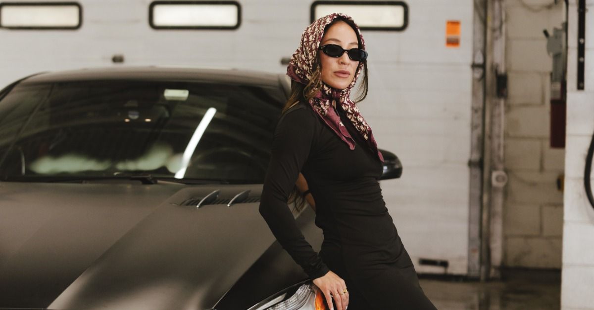 A woman wearing sunglasses and a patterned scarf leaning against a black car inside an auto service