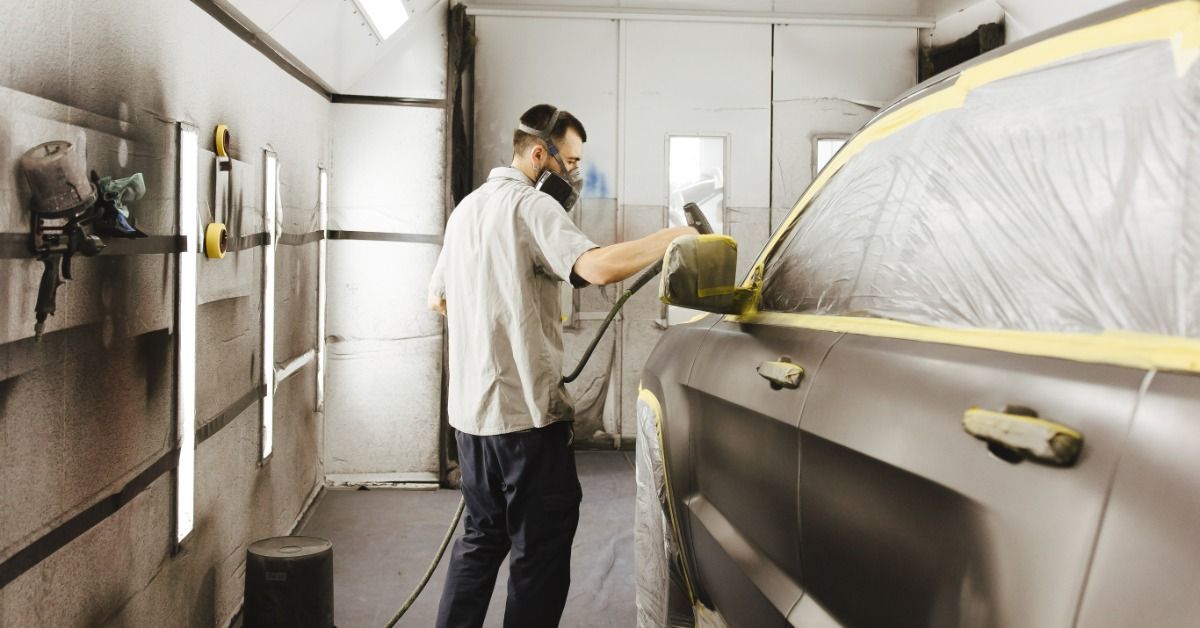 A person wearing a respirator spraying paint on a masked vehicle inside a work bay with tools mounte