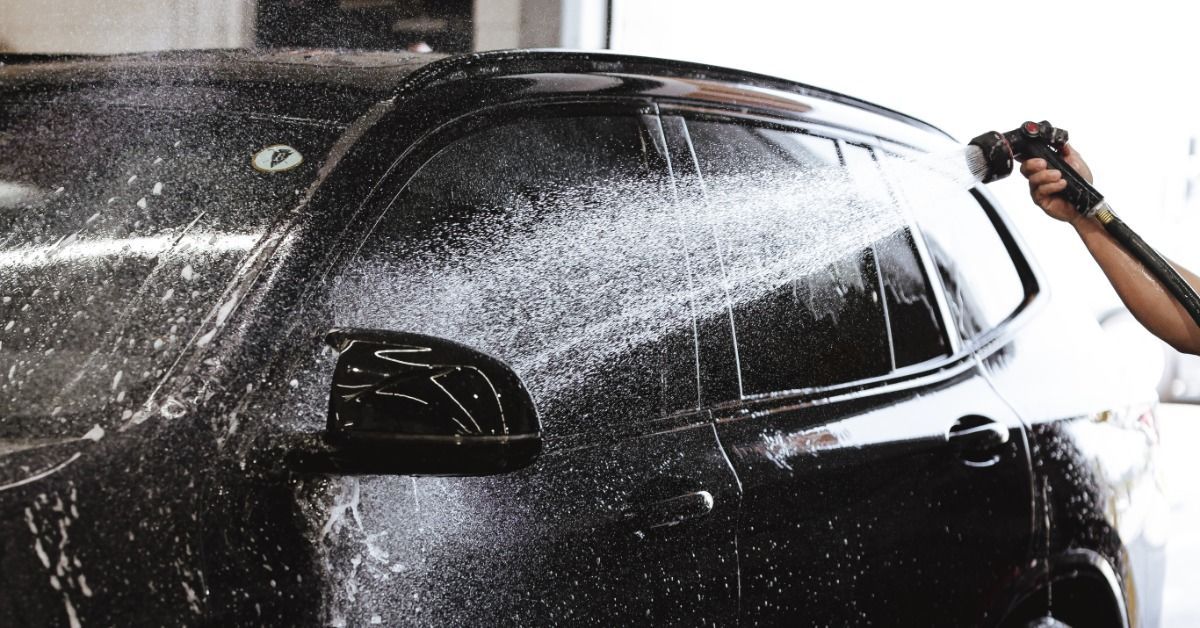 A gloved hand using a power polisher on the front of a dark vehicle beside a headlight in a work area.