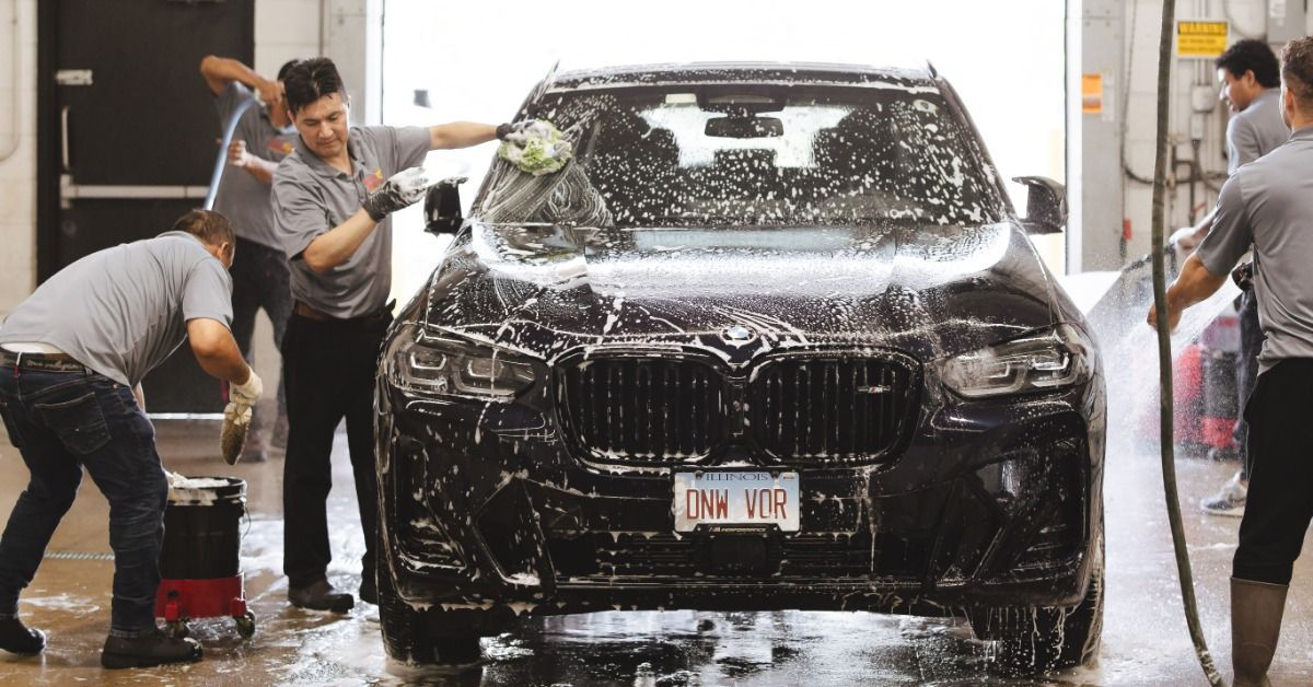 Several people washing a black vehicle covered in soap inside a garage, using sponges and water hoses.