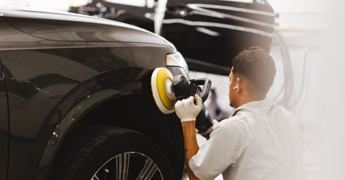 A person wearing gloves using a power polisher on the front side of a dark vehicle inside a work area.
