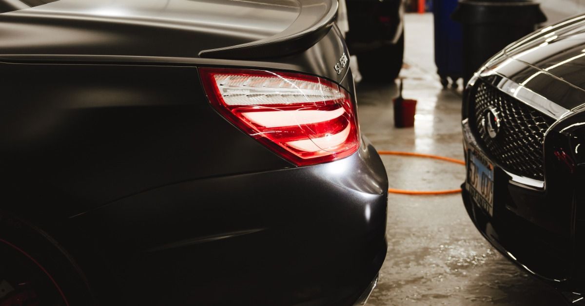 A black car with illuminated rear lights parked indoors beside another black car on a wet shop floor