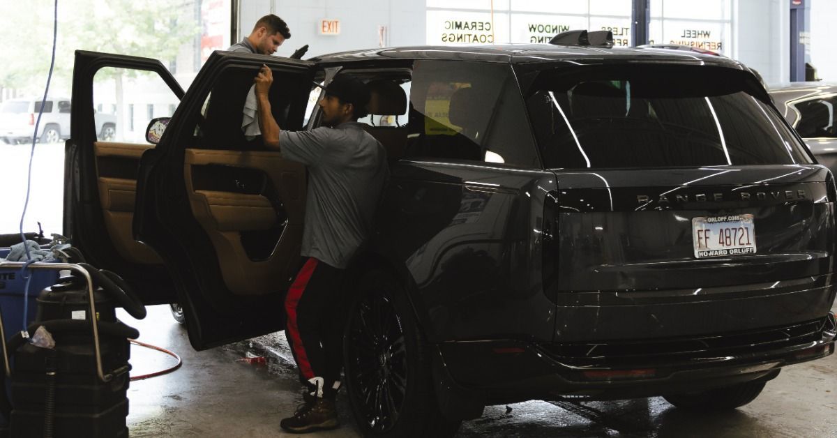 Two people standing next to a black SUV, cleaning the windows and interior. They are inside an automotive service shop.