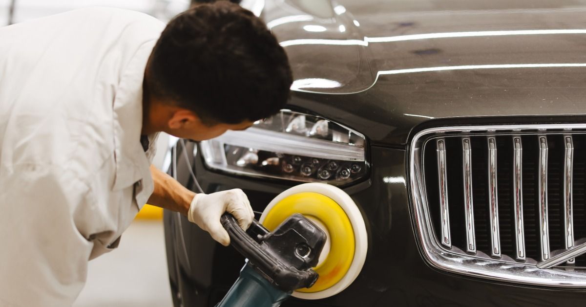 A person wearing white gloves polishing the front of a dark car with a yellow rotary buffer beside i