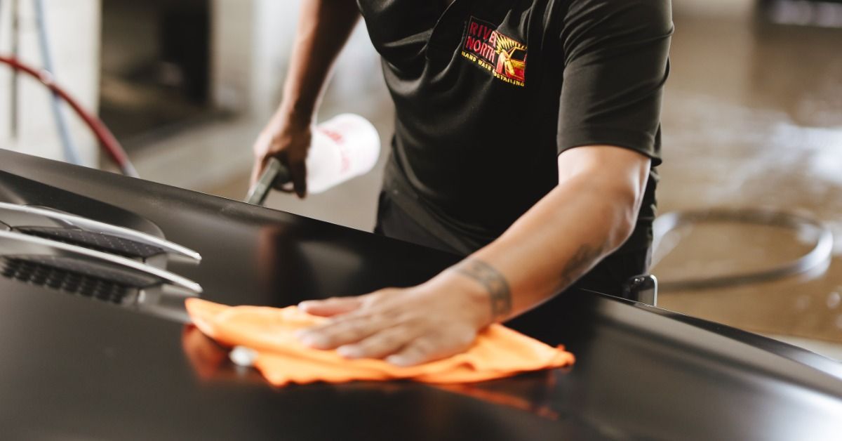 A person in a black shirt wiping the hood of a dark car with an orange cloth while holding a spray bottle.