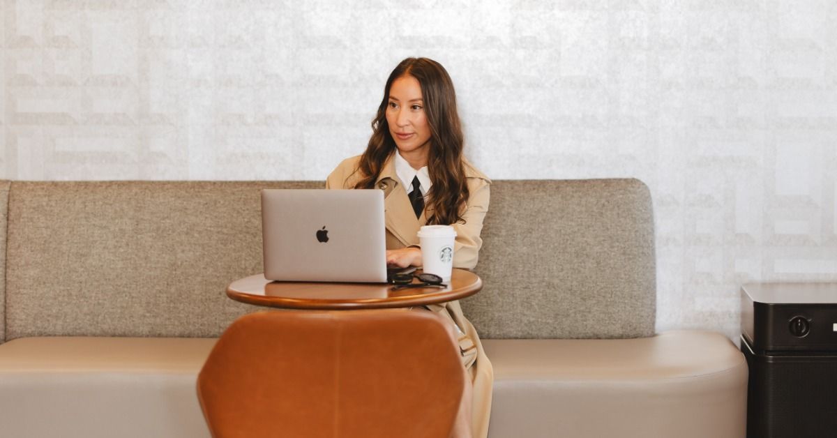 A woman with long brown hair wearing a tan coat and using a silver laptop at a round table.