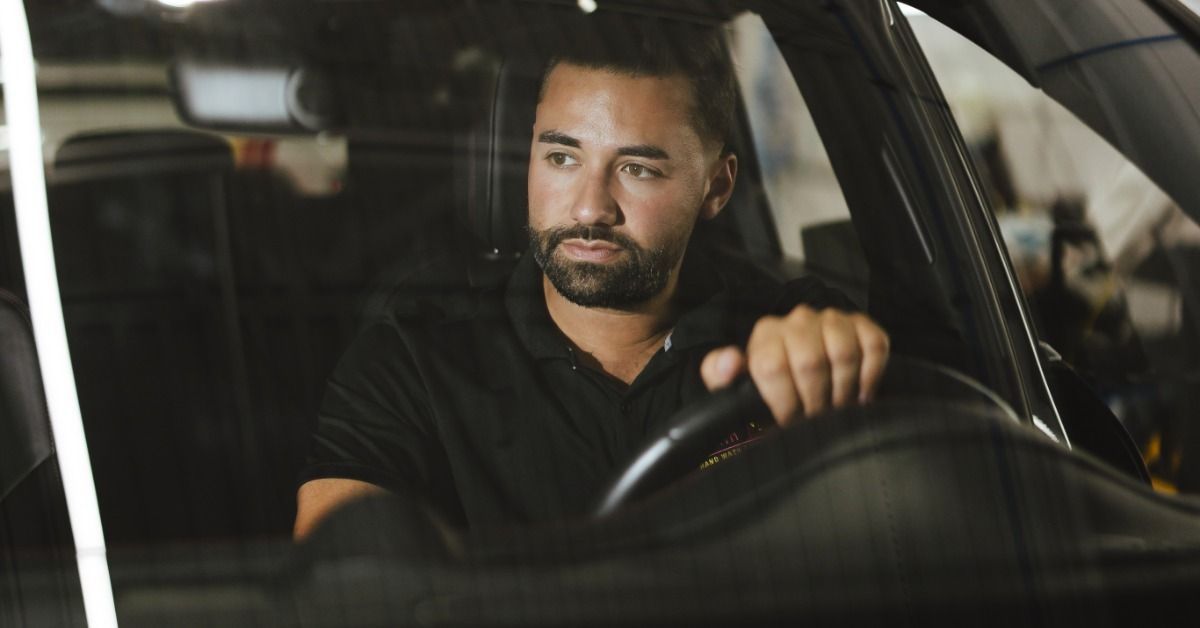A bearded man in a black polo shirt sitting behind the steering wheel of a car and looking toward the side window.