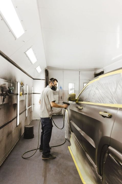 Auto body technician wearing a respirator spray paints a masked SUV inside a brightly lit automotive