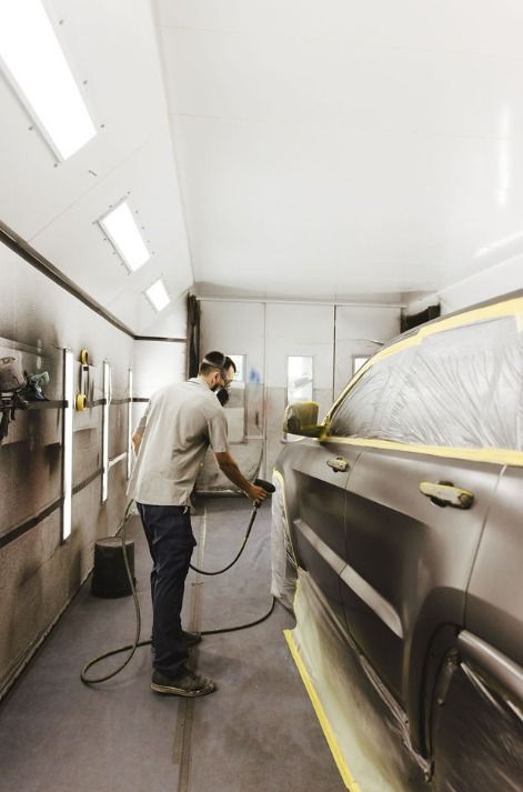Auto body technician wearing a respirator spray paints the side of a silver SUV with masked windows inside a paint booth.