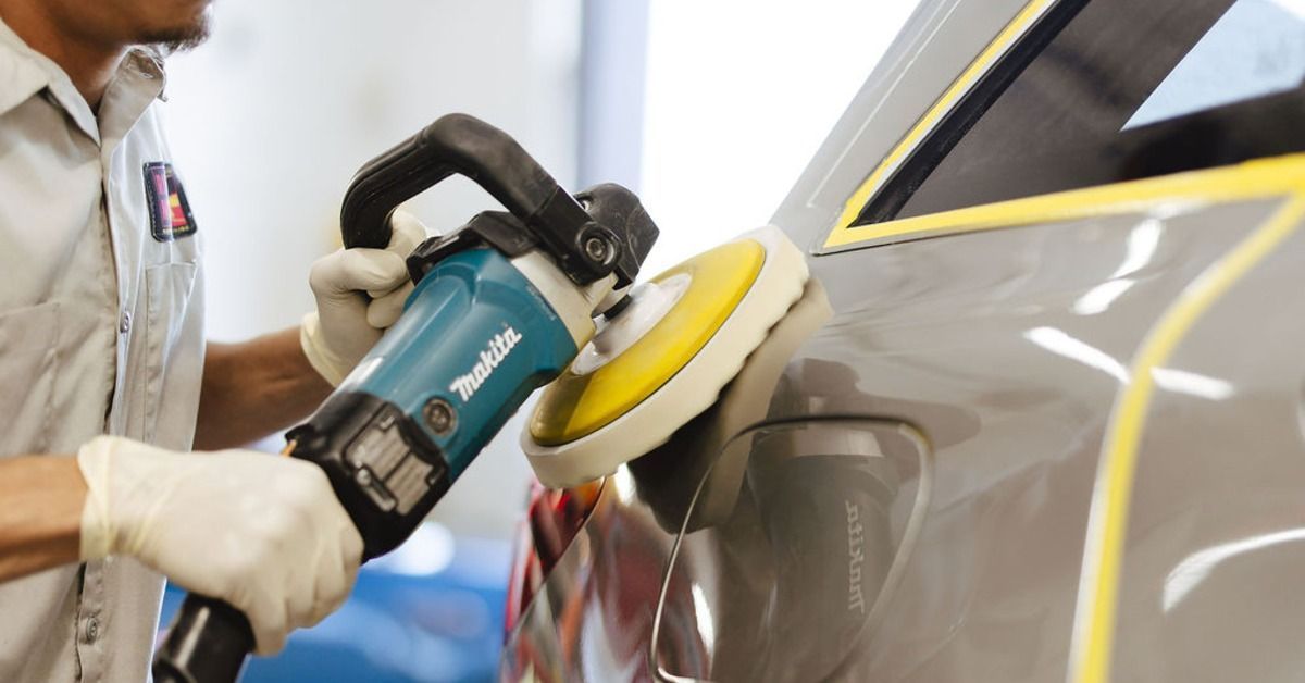 A person wearing gloves using a polisher on a gray car panel with yellow tape along the window edges