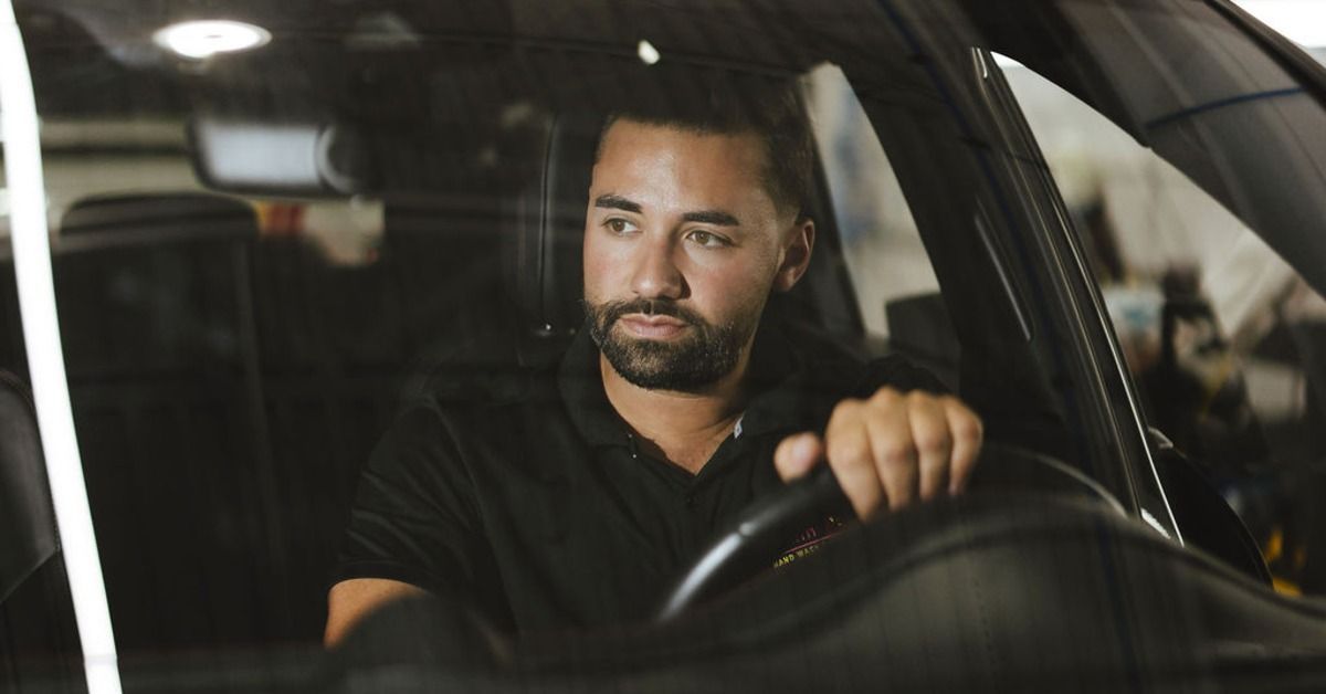 A man sitting behind the wheel of a black car inside a service bay with bright lights reflecting on the windshield.