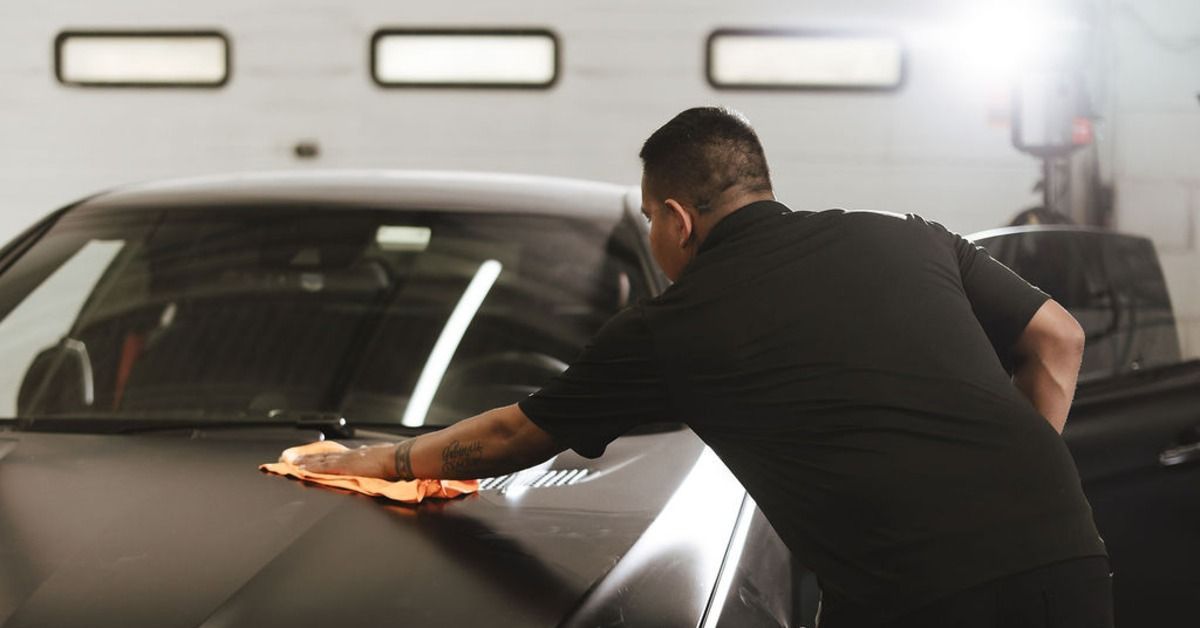 A person wiping the hood of a black car with an orange cloth inside a service bay with white garage doors.