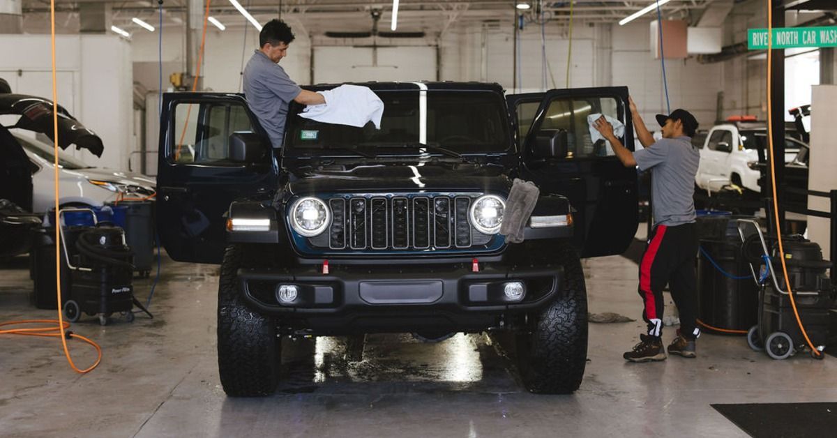 Two workers cleaning a black vehicle with all of its doors open. They are using microfiber towels inside a car wash bay.