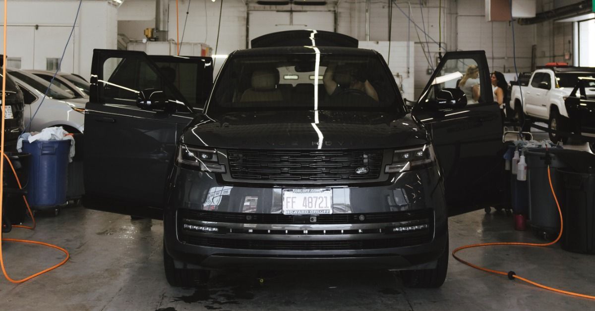 A black SUV with open doors parked inside an auto service bay with hoses, bins, other cars, and over