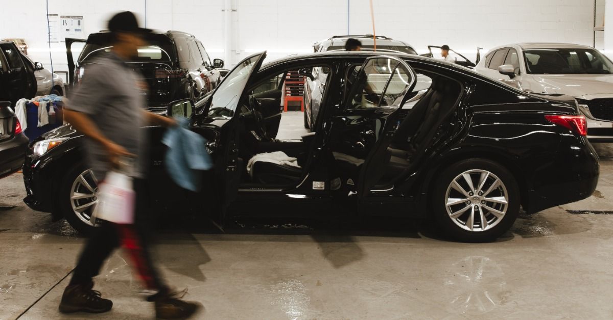 A black car with open doors parked inside an auto service bay while a worker walks past with cloths and a bottle.