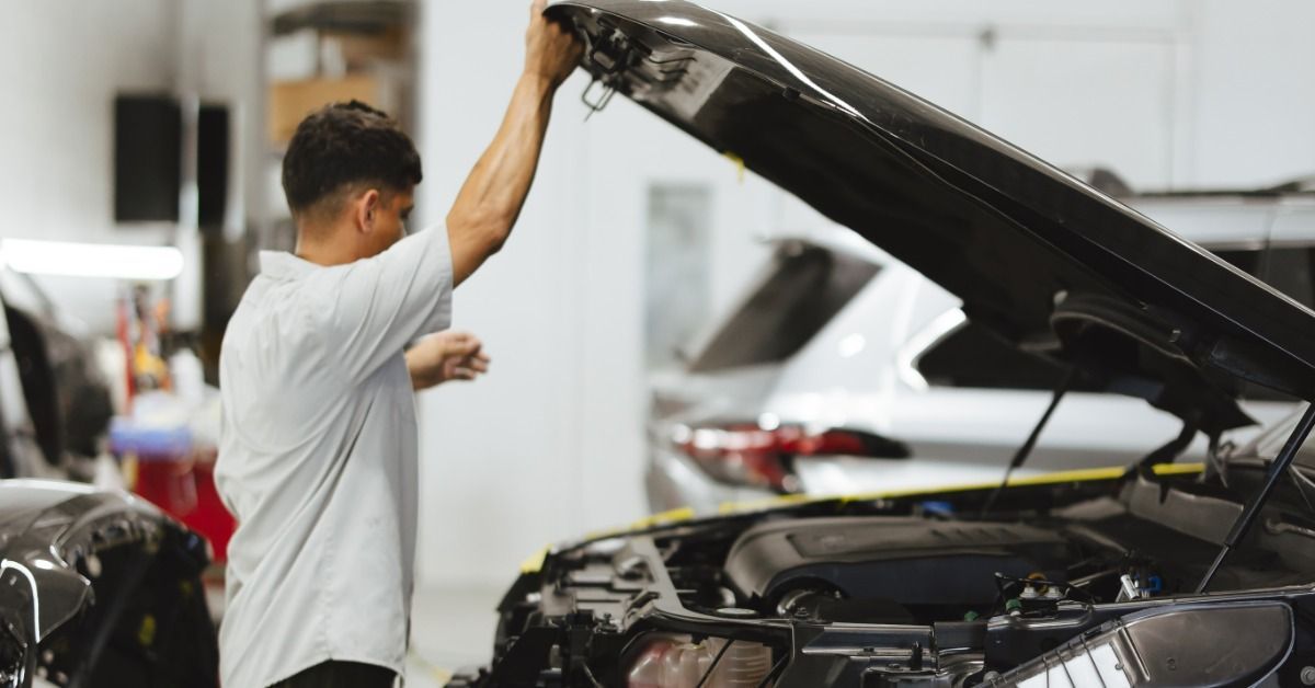A person lifting the hood of a black car inside a service bay with another vehicle and bright walls nearby.