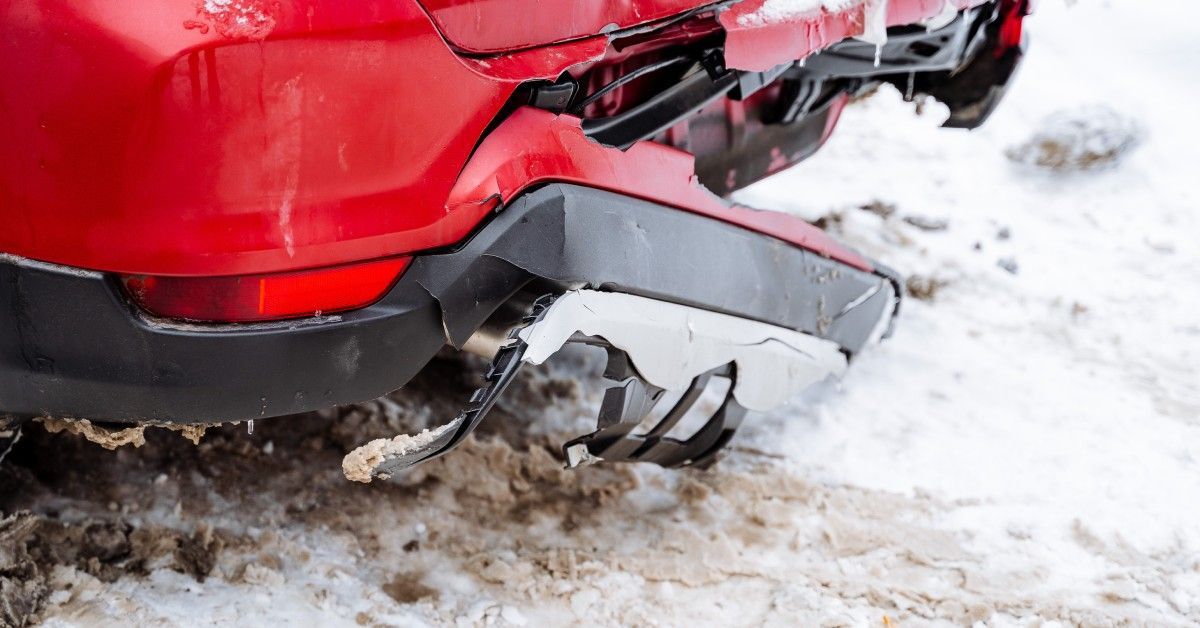 A red car has a severely damaged rear bumper. It is cracked and broken after driving in snowy conditions.