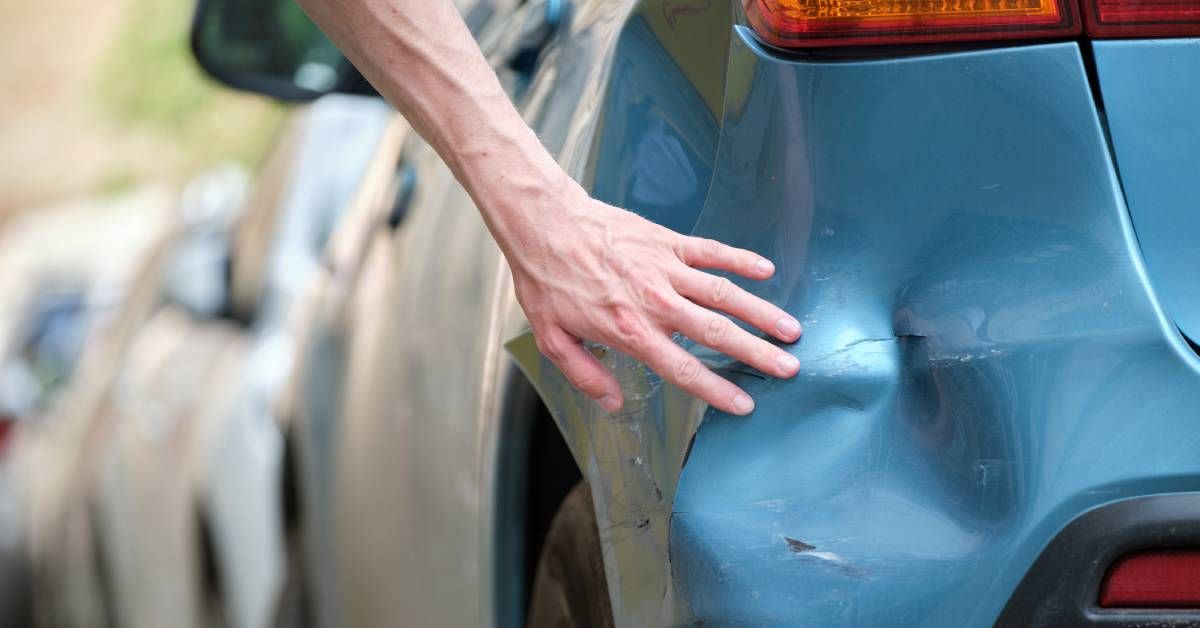 A car owner examines a large dent on their car. The dent is located at the rear of the car on the driver’s side.