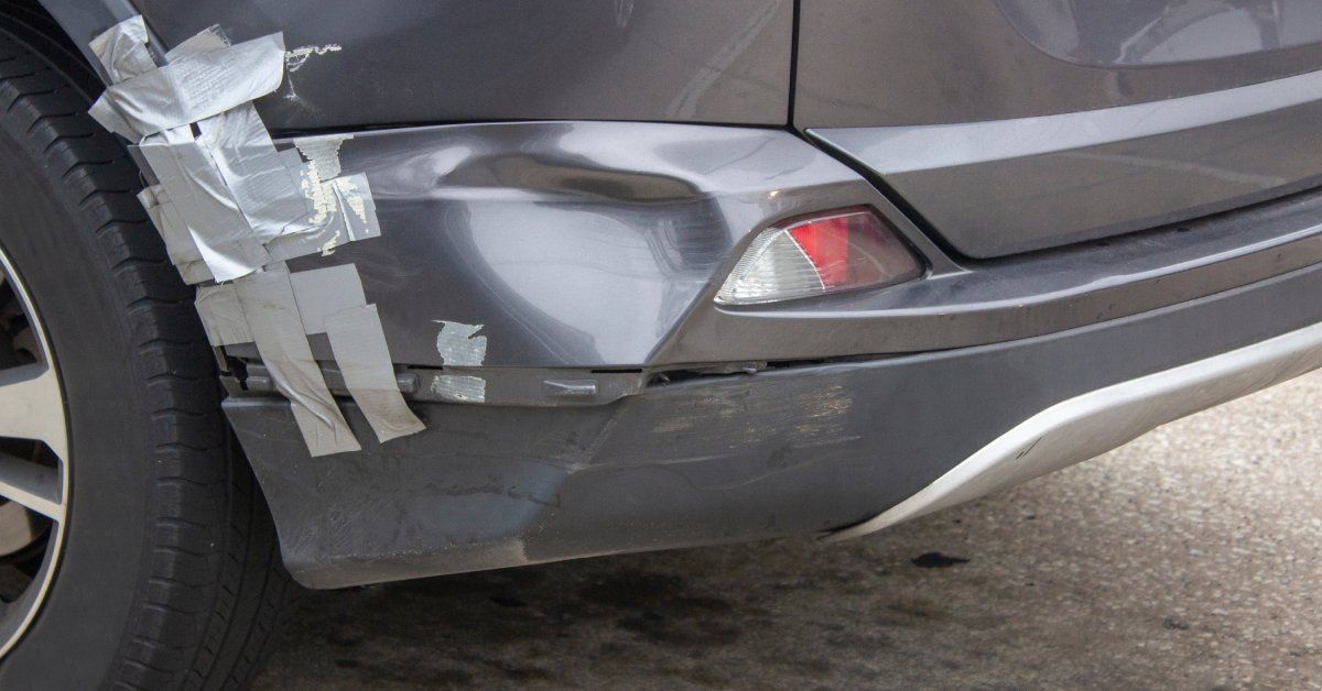 A silver car with a dent on the back bumper. There is gray duct tape holding the bumper together near the dent.