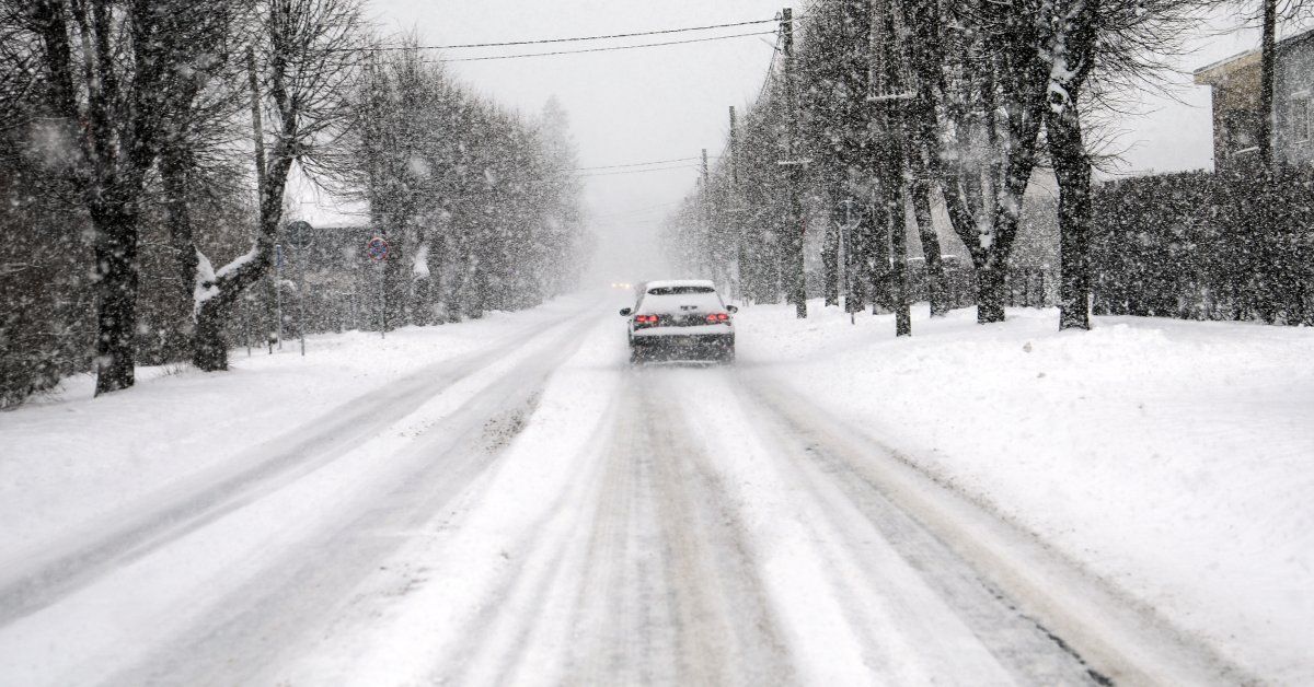 A black car driving through a snowstorm. The roads, sidewalks, and neighboring trees are covered in snow.