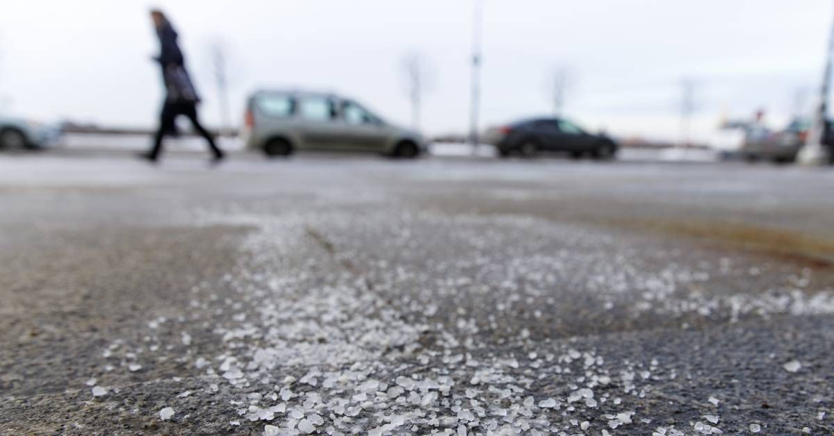Street salt particles on black asphalt. There are cars parked and a person walking on the other side of the street.