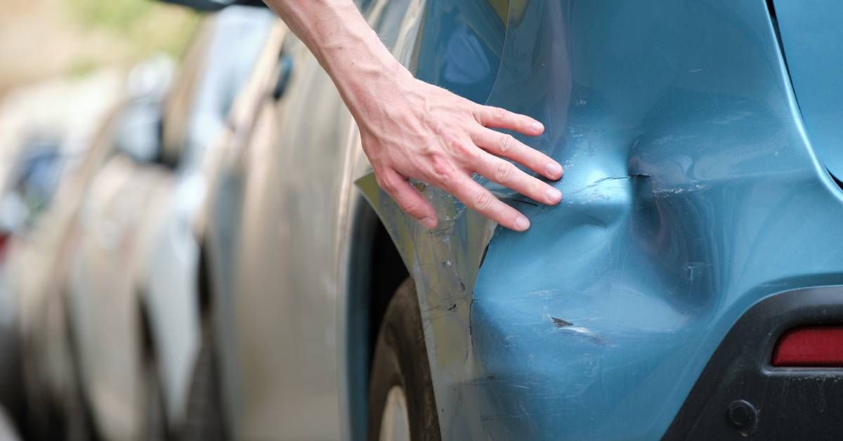 The back of a blue car with a dent on the bumper below the back left brake light. There is a hand examining the damage.
