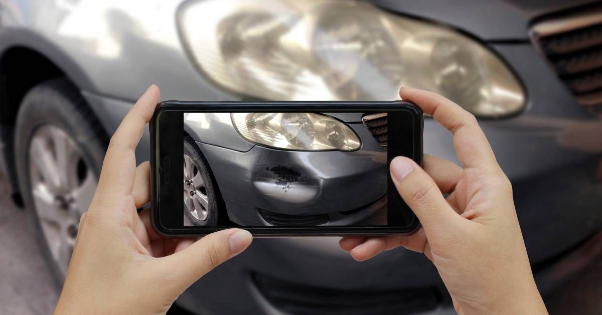 A person taking a photo of a car using their phone's camera. The car has a dent on the front bumper underneath the headlight.